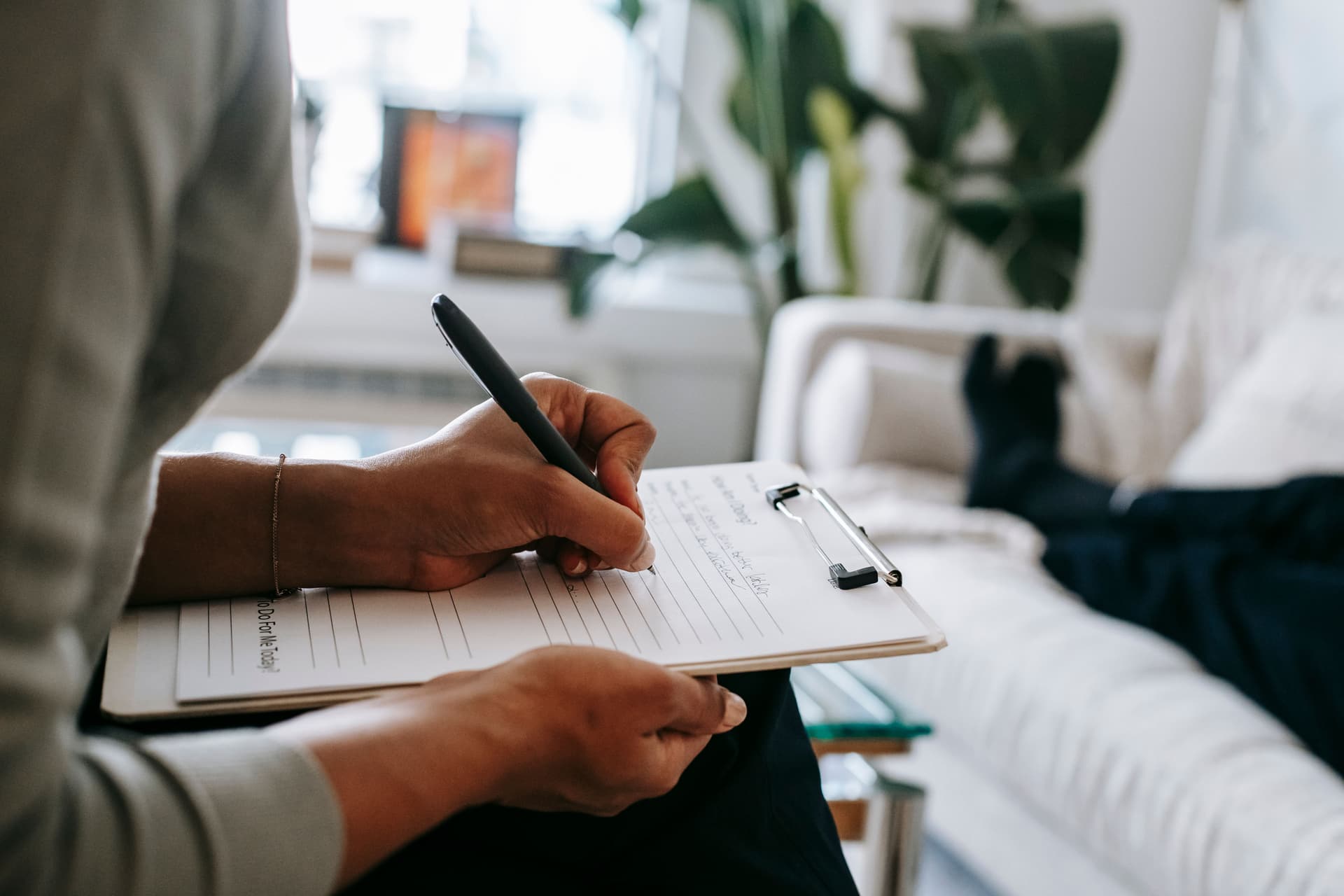 A therapist writes on a clipboard during a session while a client rests on a couch nearby.